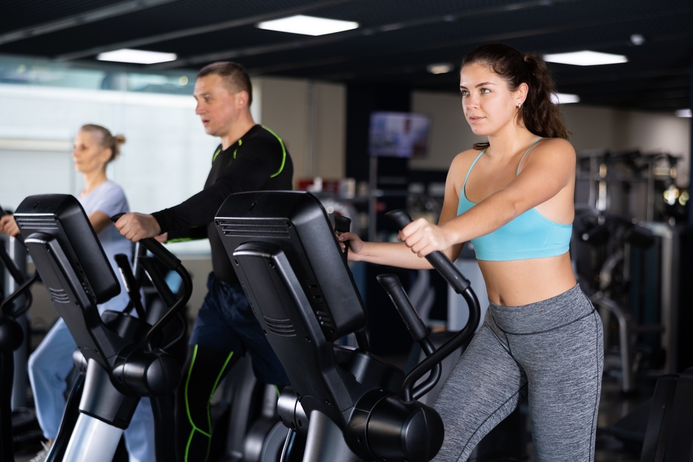 Motivated sporty young girl doing cardio training, exercising on elliptical trainer in gym..
