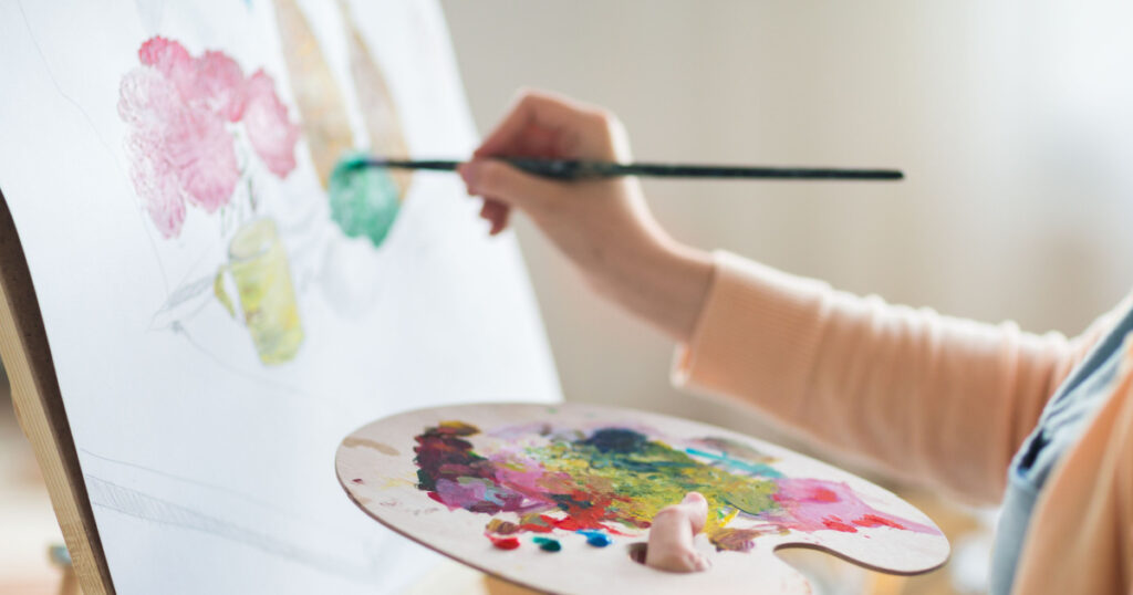art, creativity and people concept - close up of artist with palette and brush painting still life on paper at studio