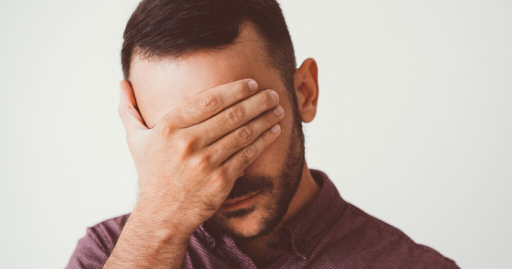 Closeup of young man covering eyes with hand. Depression concept. Isolated front view on grey background.