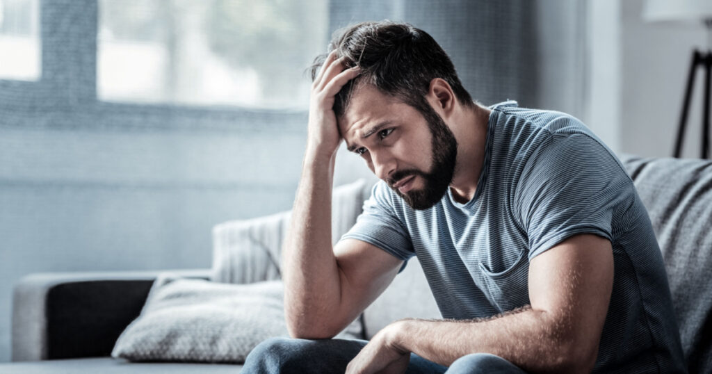 Unpleasant pain. Sad unhappy handsome man sitting on the sofa and holding his forehead while having headache