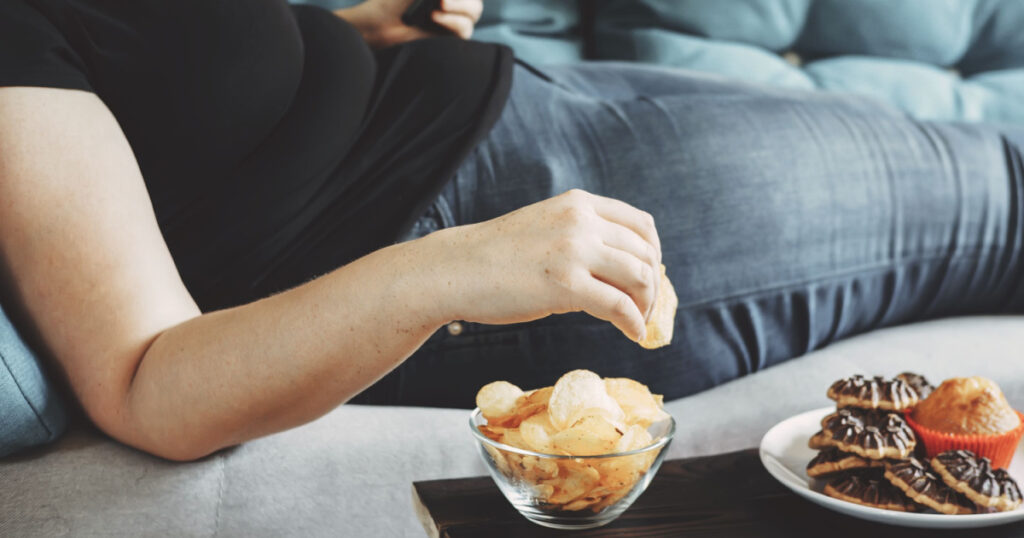 g, sedentary lifestyle, compulsive overeating. Obese woman laying on sofa with smartphone eating chips