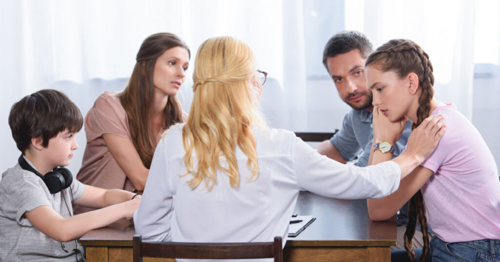 rear view of female counselor cheering up upset teenage girl on family therapy session in office