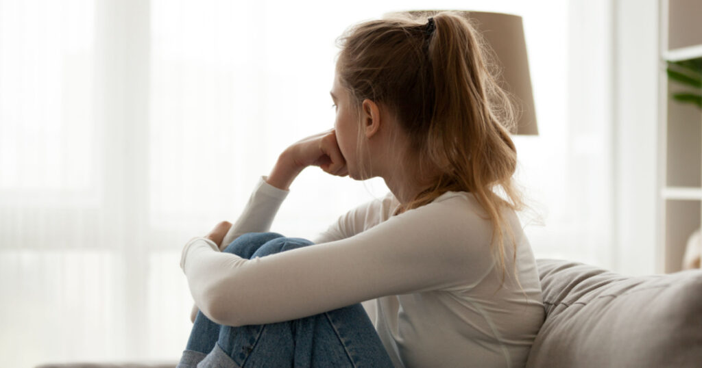 Side view young woman looking away at window sitting on couch at home. Frustrated confused female feels unhappy problem in personal life quarrel break up with boyfriend or unexpected pregnancy concept
