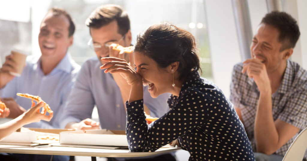 Indian woman laughing at funny joke eating pizza with diverse coworkers in office, friendly work team enjoying positive emotions and lunch together, happy colleagues staff group having fun at break