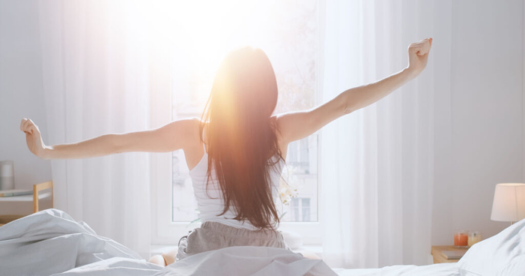 Beautiful Brunette is Waking up in the Morning, Stretches in the Bed, Sun Shines on Her From the Big Window. Happy Young Girl Greets New Day with Warm Sunlight Flare.