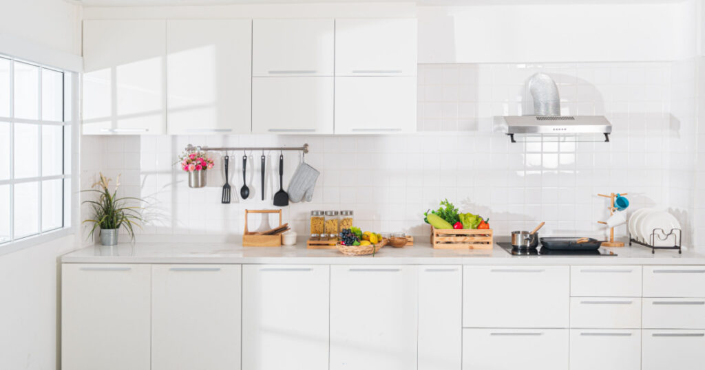 Modern white kitchen with counter and white details, minimalist interior with sunlight in daytime. Full set of kitchen equipment, pan, pot, electric hob, flipper, vegetable, fruit.