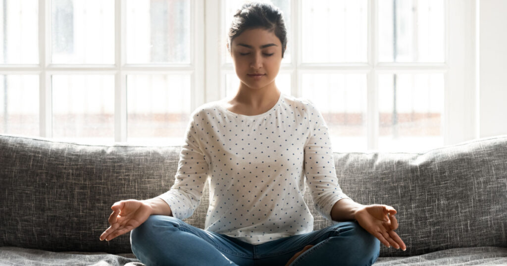 Full length mindful young indian woman making mudra gesture, sitting in lotus position on comfortable couch at home. Peaceful millennial girl deeply meditating, doing breathing yoga exercises alone.