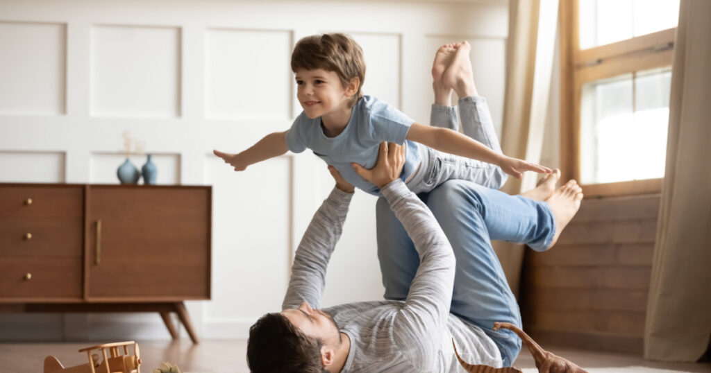 Joyful young man father lying on carpet floor, lifting excited happy little child son at home. Full length carefree two generations family having fun, practicing acroyoga in pair in living room.