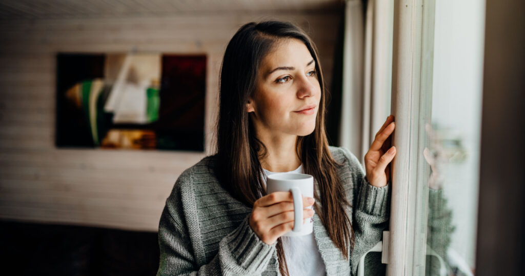 Young woman spending free time home.Self care,staying home.Enjoying view,gazing through to the window.Quarantined person indoors.Serene mornings.Avoiding social contact.