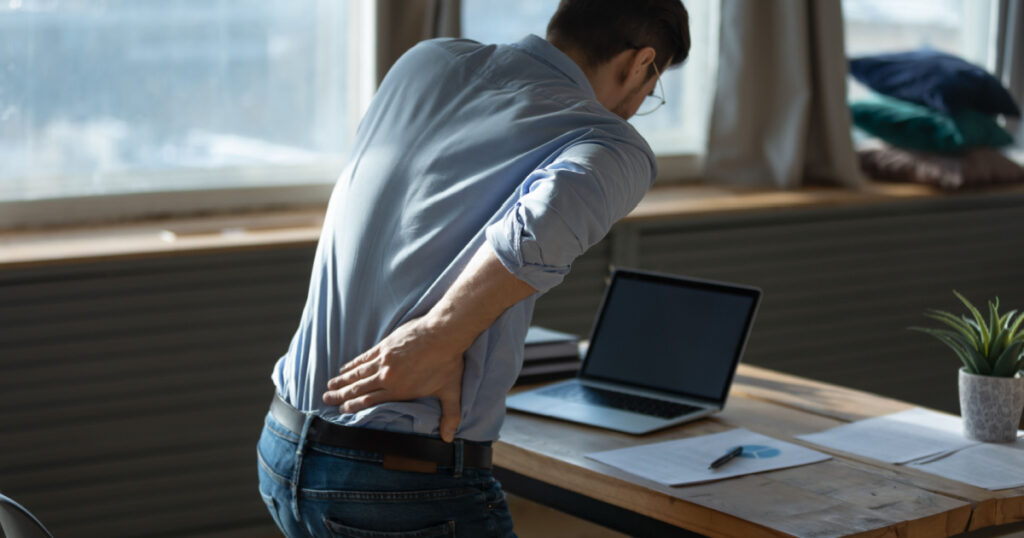 Rear view young man suffering from sudden backache, getting out of uncomfortable chair at workplace, touching lower back, unhealthy businessman student office worker feeling discomfort