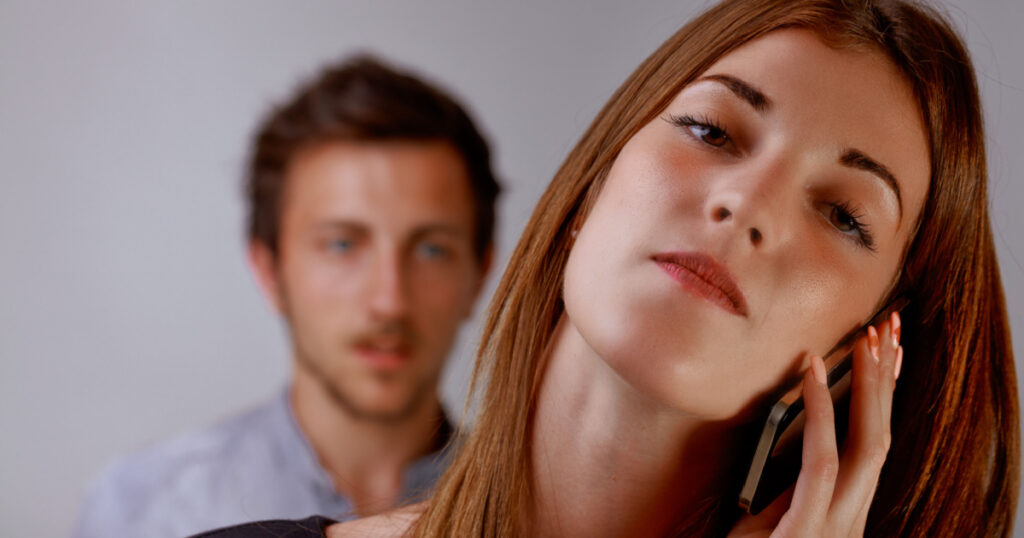 concept of narcissistic woman cheating on man who loves her: young attractive and self-confident woman on cell phone while man behind her looks sad