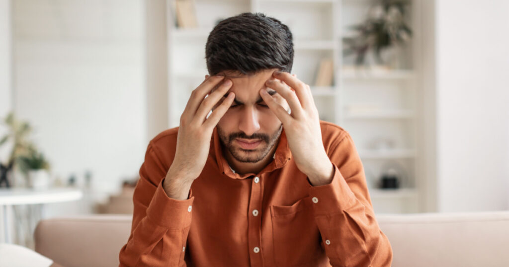 Young Middle Eastern guy suffering from headache after hard working day, sitting on couch at home, massaging head temples, having strong migraine, feeling tired, upset and stressed, copy space