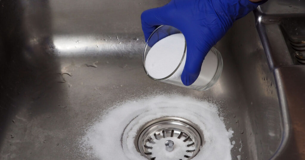 Gloved hand putting baking soda on drain in kitchen sink from glass jar. Close up. Eco friendly house cleaning concept.