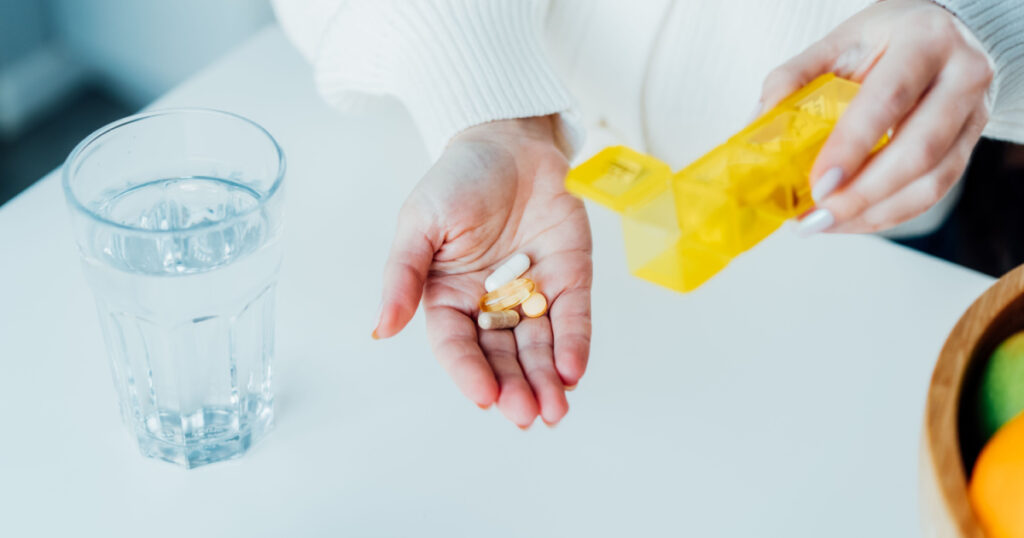 Close-up of woman's hand holding pills and pill organizer at home kitchen with water glass. Daily intake of antioxidant diet, vitamin supplements for beauty, skin, hair, healthcare, drugs concept.