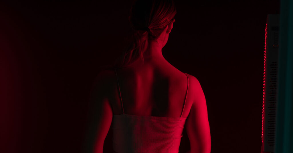 Rearview of a woman receiving red light therapy on her back. Woman standing next to a red light device in a cosmetic spa. Anti-aging skincare treatment.