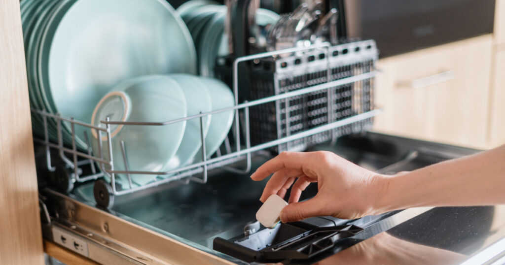 Close up view of female hand inserting dishwasher tablet into open built-in dishwasher machine with utensils inside in modern home kitchen. Household, housekeeping, domestic concept