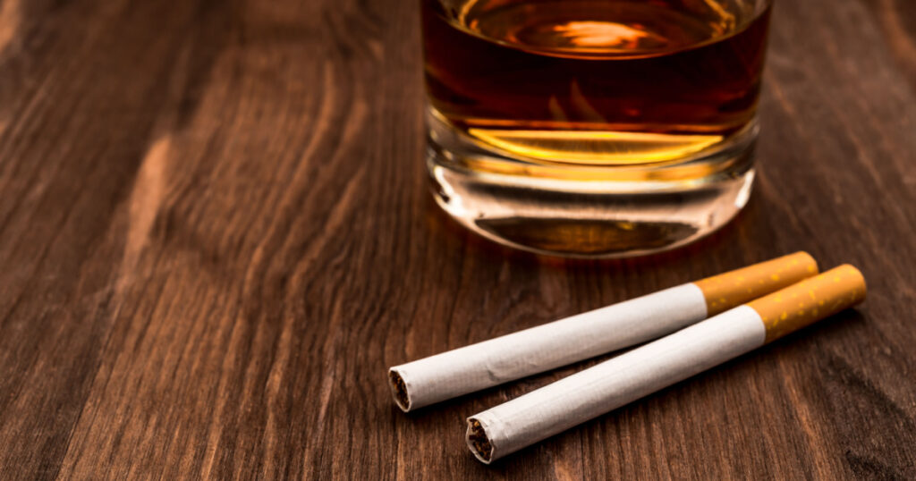 Glass of whiskey and two cigarettes on a wooden table. Focus on the cigarettes