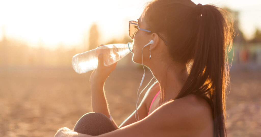 Beautiful fitness athlete woman drinking water after work out exercising on sunset evening summer in beach outdoor portrait