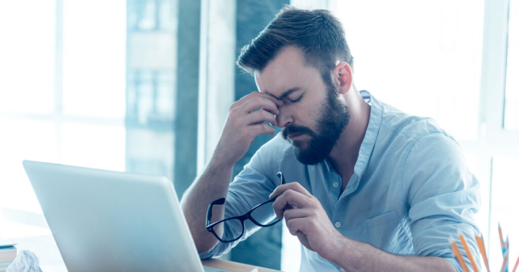 Feeling exhausted. Frustrated young beard man massaging his nose and keeping eyes closed while sitting at his working place in office
