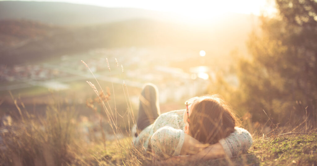 Carefree happy woman lying on green grass meadow on top of mountain edge cliff enjoying sun on her face.Enjoying nature sunset.Freedom.Enjoyment.Relaxing in mountains at sunrise.Sunshine.Daydreaming