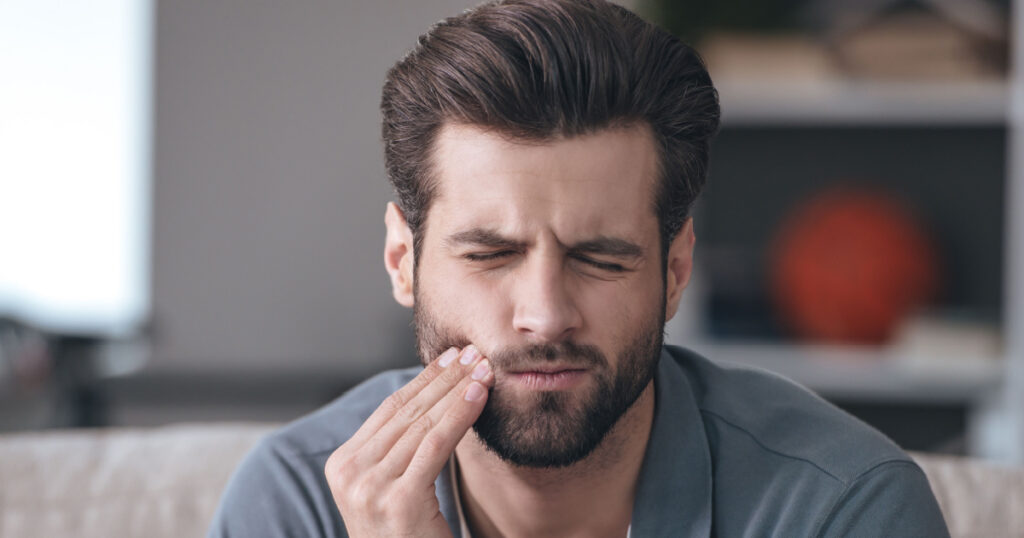 Toothache. Frustrated young man touching his cheek and keeping eyes closed while sitting on the couch at home