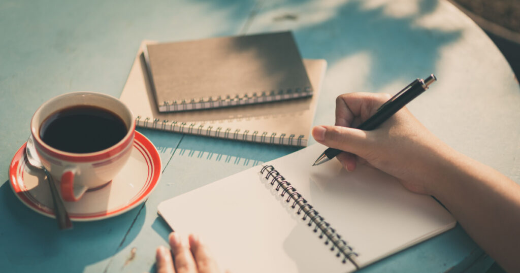 Woman right hand writing journal on small notebook at outdoor area in cafe with morning scene and vintage filter effect