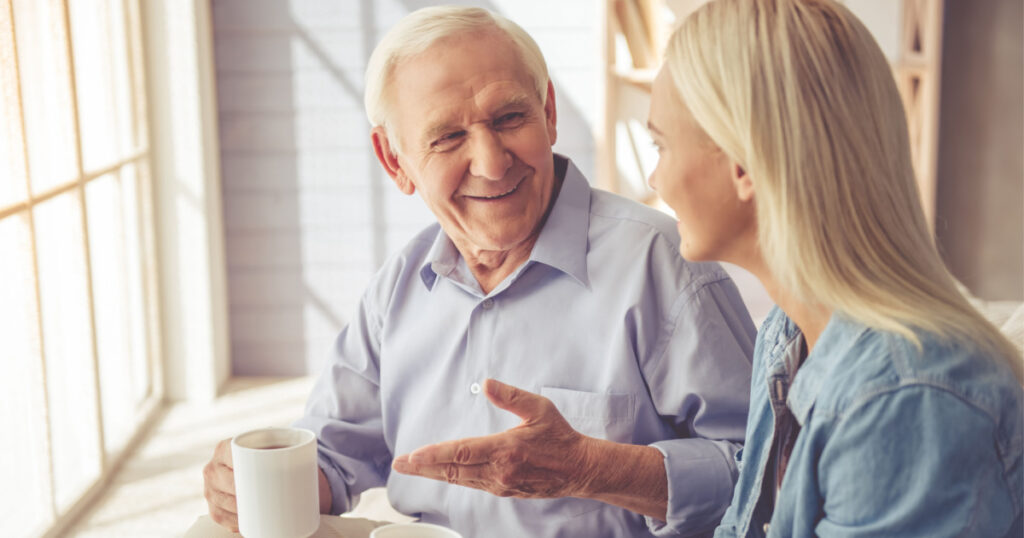 Handsome old man and beautiful young girl are drinking tea, talking and smiling while sitting on couch at home
