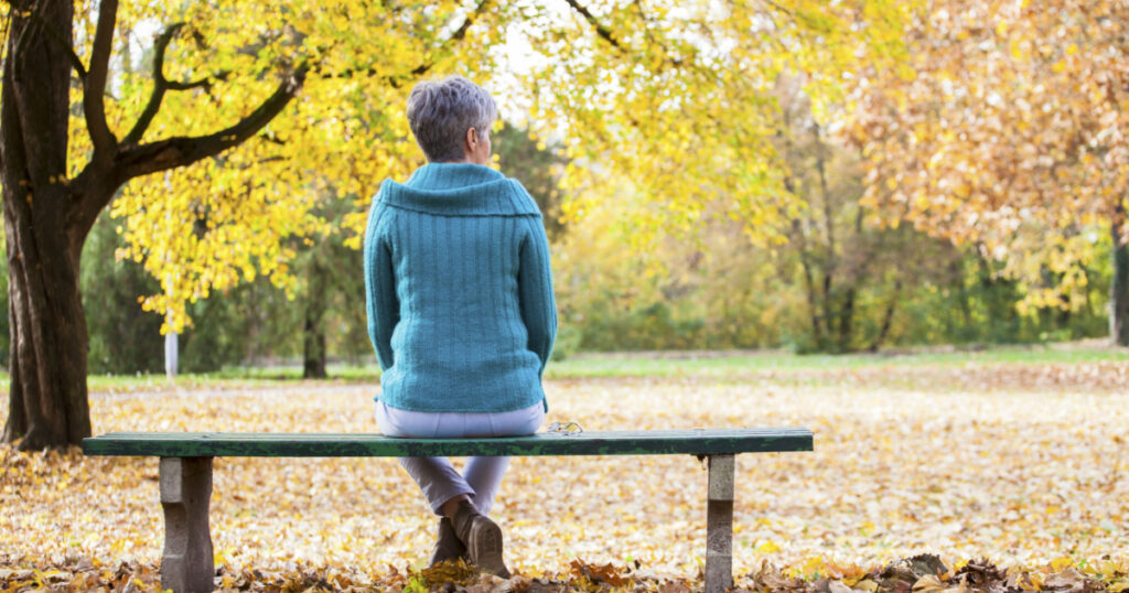 Depressed and sad old woman on bench in autumn park, from back angle