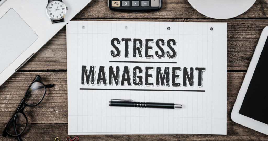 Stress Management statement on paper note pad. Office desk with electronic devices and computer, wood table from above, concept image for blog title or header image. Aged vintage color look.