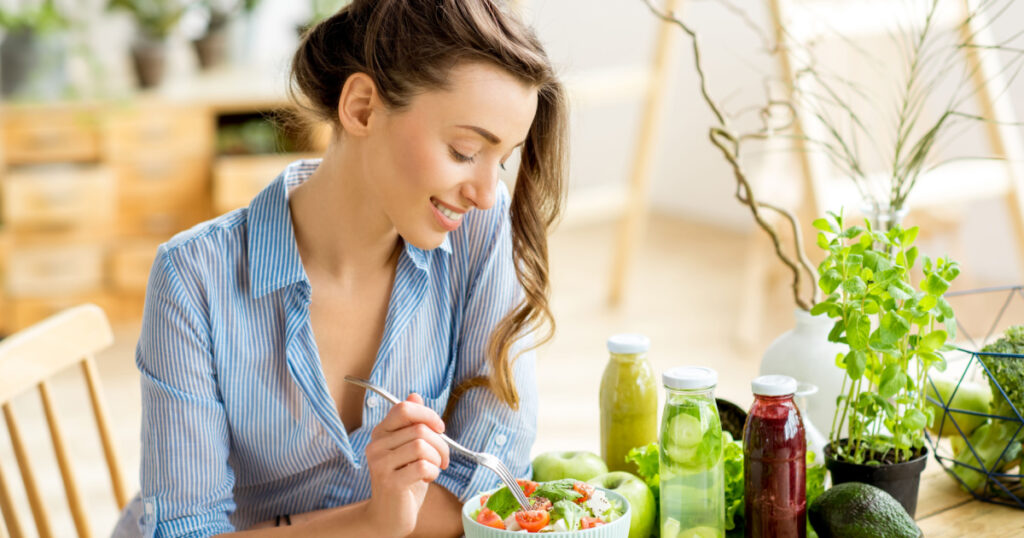 Young and happy woman eating healthy salad sitting on the table with green fresh ingredients indoors