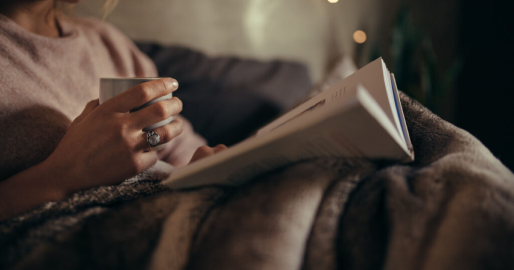 Cropped shot of female hands with book and coffee. Female reading book on bed at night.