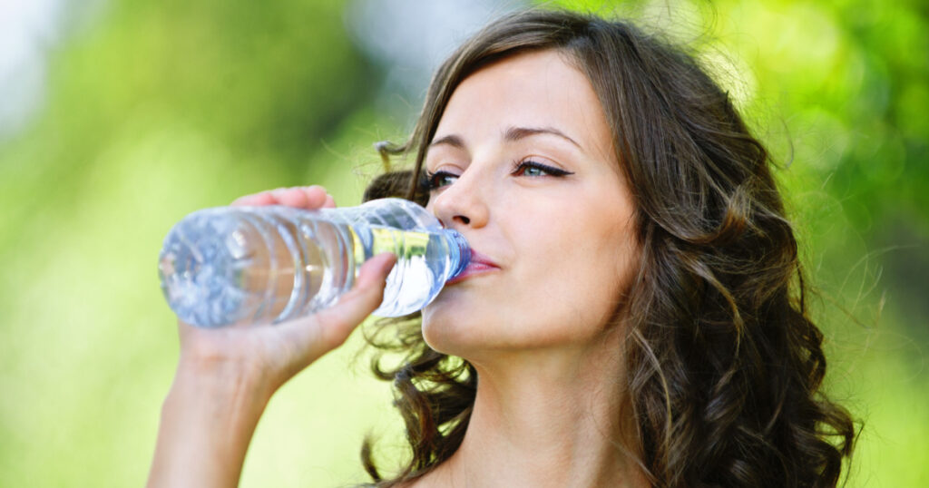 Portrait of young beautiful dark-haired woman wearing blue t-shirt drinking water at summer green park.