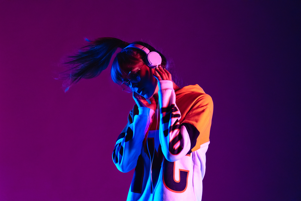 Stylish fashion teenager model wearing hoodie and headphones listening dj music dancing in purple neon lights. Young teen girl enjoy cool music 90s party mix in violet studio background. Copy space.