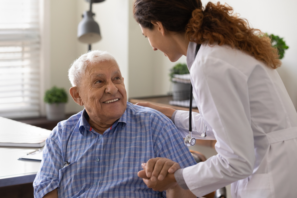 Close up caring nurse holding smiling mature patient hand at meeting in hospital, doctor caregiver wearing uniform comforting and supporting senior man, good news about treatment, empathy and care
