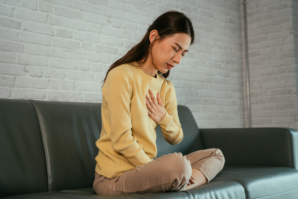 Asian young woman feeling discomfort as suffering from heartburn holding chest with closed eyes and sitting with folded legs on couch at home
