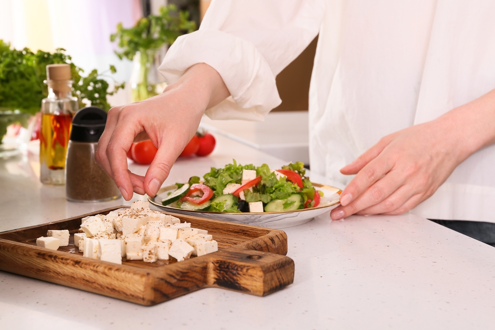 Woman making Greek salad with cut feta cheese on table in kitchen
