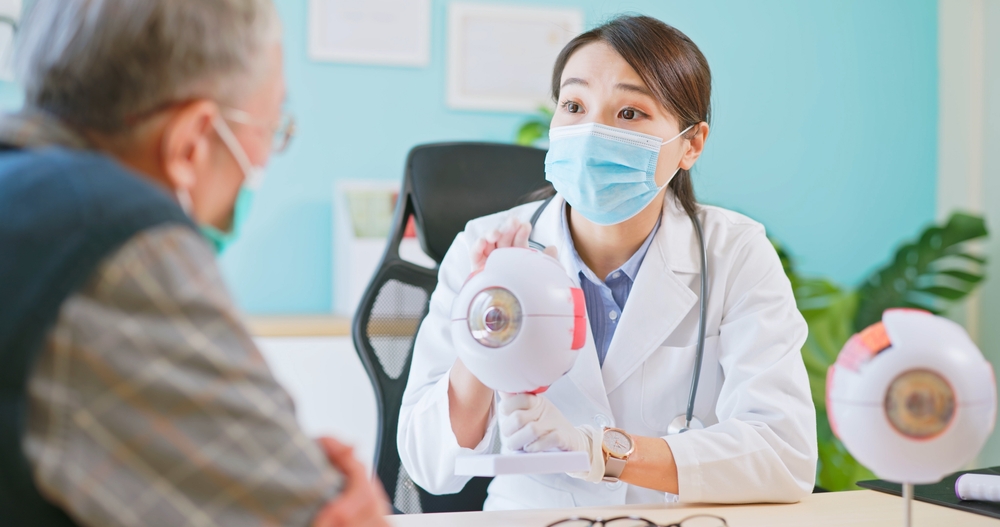 asian female doctor wearing face mask is showing eyeball model and explaining about eyes disease to elder senior man patient in hospital