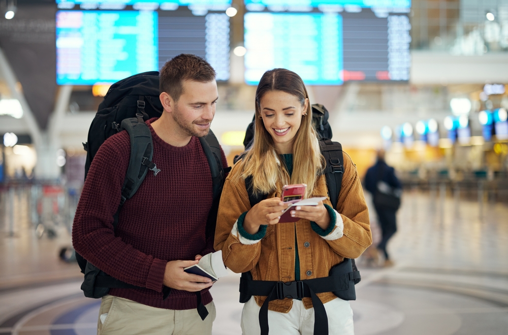Happy couple, airport and phone with ticket, travel app and adventure with excited face, conversation and smile. Man, woman and smartphone for digital booking of hotel, taxi or bus for transportation
