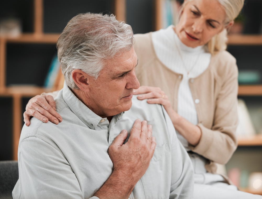 Heart attack, pain and senior couple at home with cardiology problem or stroke. House, cardiac arrest and retirement of a man with emergency and health issue in a living room with female support
