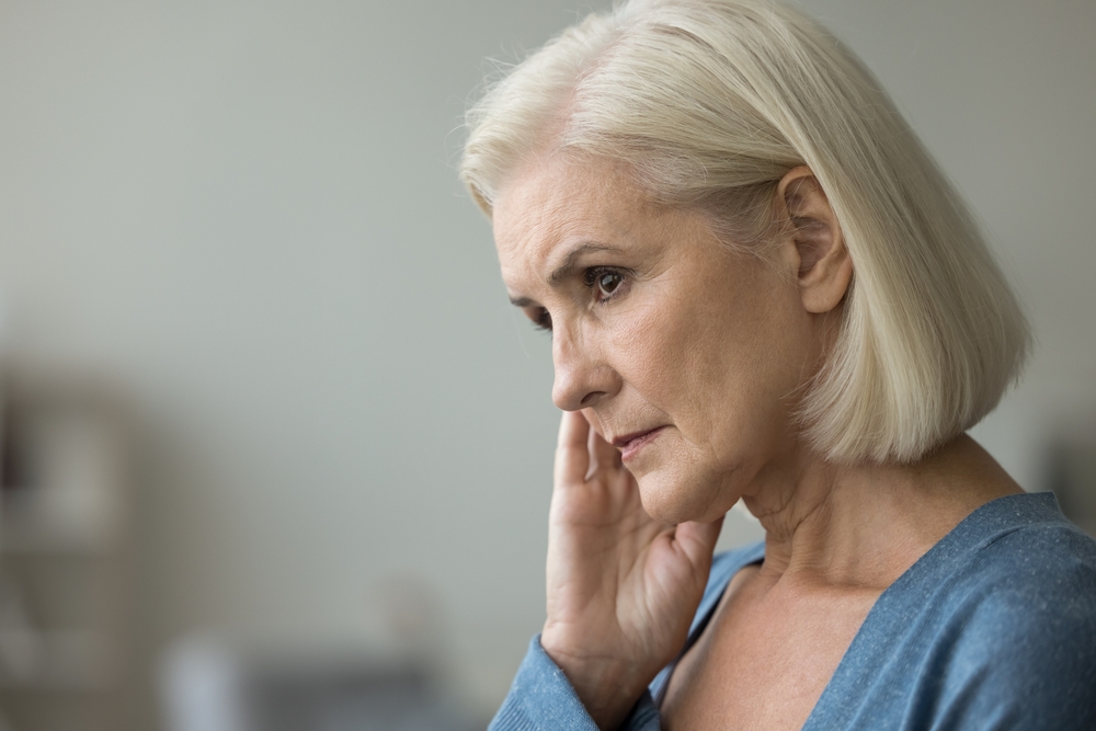 Serious thoughtful blonde elderly woman worried about health problems, thinking over retirement, touching dry facial skin, face, looking away, feeling depressed. Side casual portrait