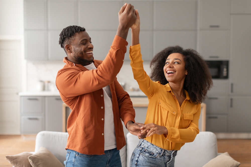Cheerful millennial black spouses having fun together dancing to music in modern kitchen interior, smiling to each other. Couple having date or party. Relationship and romance concept