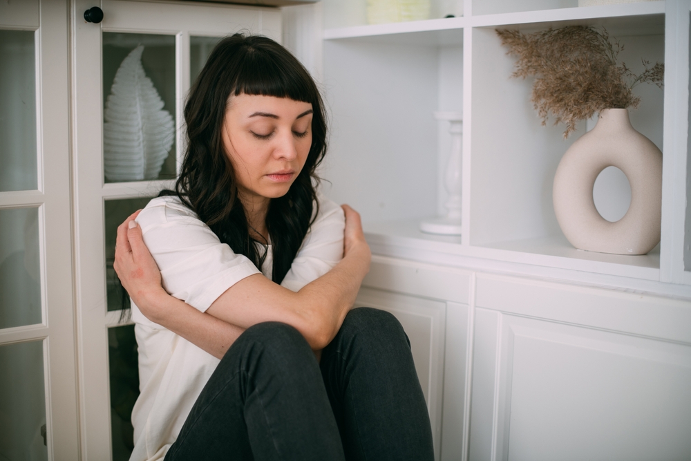 A young woman sadly sits in a corner and hugs herself in a bright room. The concept of the inner child and the protection of the inner world. A beautiful sad girl sits on the floor in secluded corner
