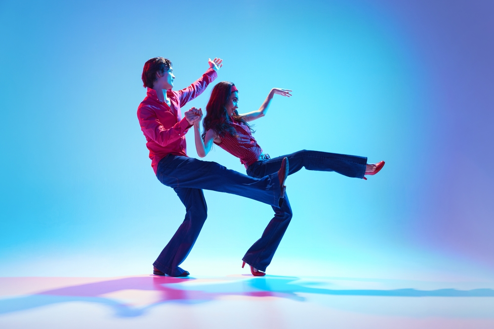 Happy, positive young man and woman in retro clothes dancing boogie woogie retro dance against blue background in neon light. Concept of hobby, dance class, party, 50s, 60s culture, youth