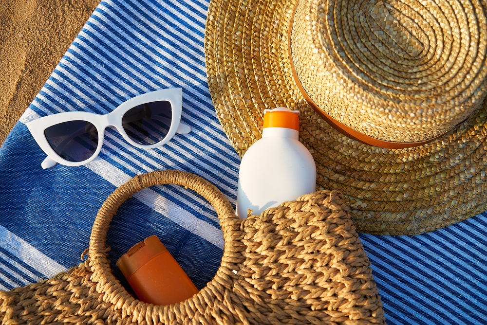 Beach scene with straw, white sunglasses, sunscreen in wicker bag on blue towel. Summer essentials for sun protection on sandy shore by ocean. Relaxing day at beach necessary sunbathing.