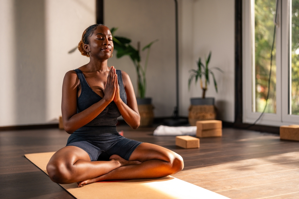 Young black girl practicing yoga indoors, sitting cross-legged on a yoga mat with her hands in a prayer position. She appears calm and focused, with plants and yoga blocks in the background.
