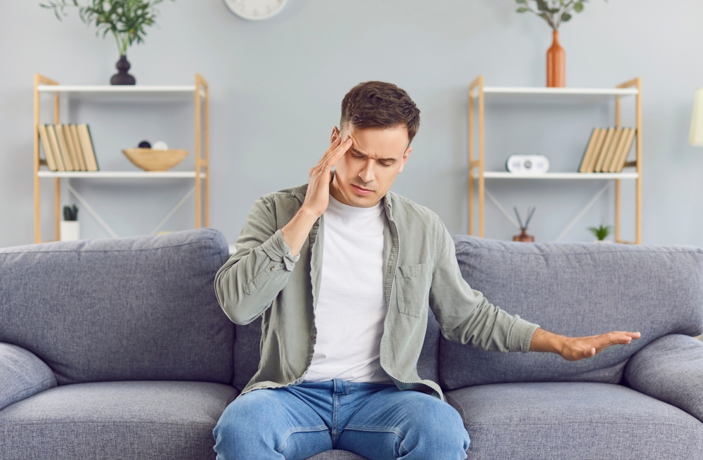 Unhealthy man suffering from migraines or dizziness sits on the couch at home, feels pain in his head. He is stressed, experiencing vertigo or headache, worrying about work or relationship problems.
