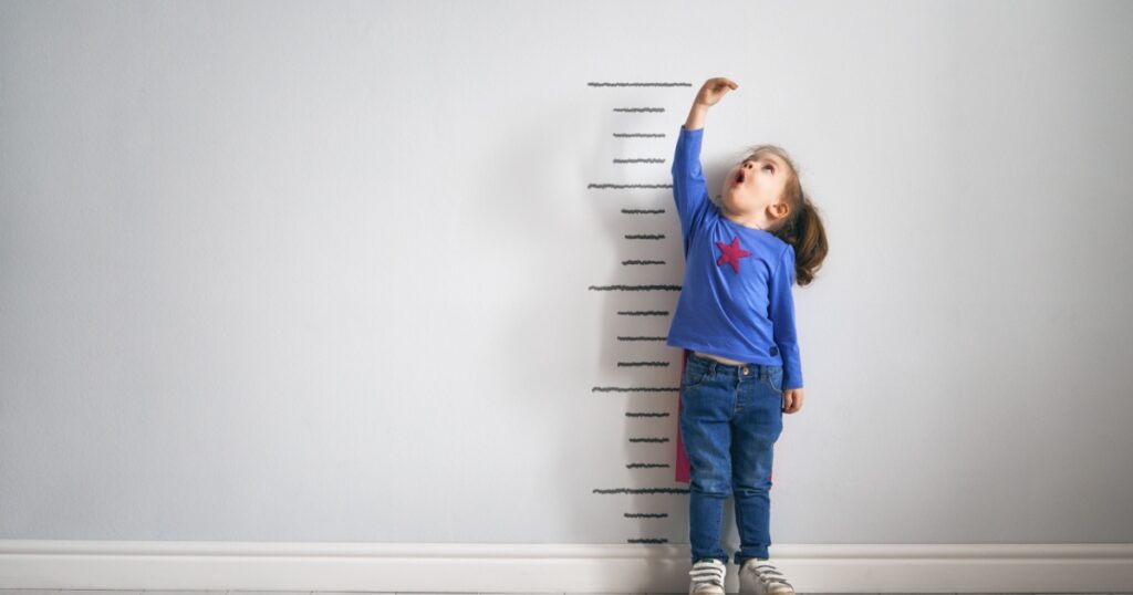 Little child is playing superhero. Kid is measuring the growth on the background of wall. Girl power concept.