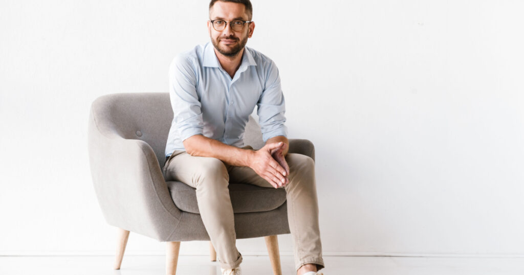Horizontal portrait of attractive caucasian guy wearing stylish formal clothing sitting in armchair and looking at camera isolated over white background