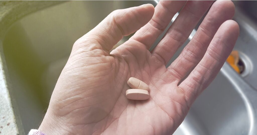 Old hand holding pills over a sink.