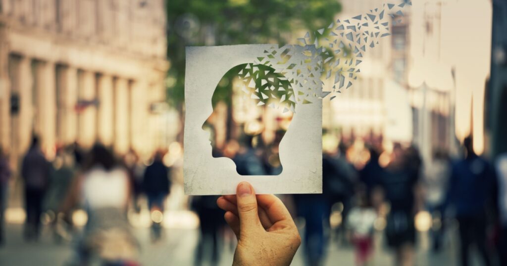 Hand holding a paper sheet with human head icon broken into pieces over a crowded street background. Concept of memory loss and dementia disease. Alzheimer's losing brain and memory function.
